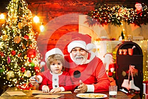Funny boy in Santa hat writes letter with Santa near christmas tree. Portrait of cheerful dreamy granny Santa and