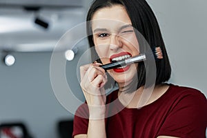 Funny brunette female with red lips bites a brush making make-up. Happy young woman making herself makeup with a brush with