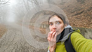 Funny brunette girl takes a selfie depicting fright on the background of a mysterious foggy road. Independent walk