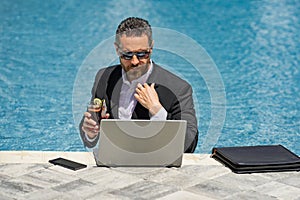 Funny businessman in suit working with computer on the sea beach. Summer holiday, outdoor and business travel concept