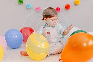 Funny Caucasian baby boy celebrating first birthday. Child kid toddler sitting on floor with colorful balloons. Celebration of