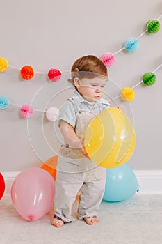 Funny Caucasian baby boy celebrating his first birthday. Excited child kid toddler playing with colorful balloons. Celebration of