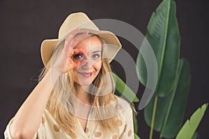 Funny caucasian blonde young girl standing in front of a big-leaf plant in a hat doing ok gesture with hand smiling, eye