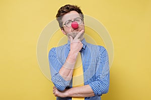 Funny caucasian man in red clown nose and yellow tie thinking with a hand in his chin