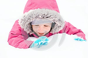 Funny Caucasian smiling girl in warm winter clothes pink jacket playing with snow. Cute child lying on ground during cold winter