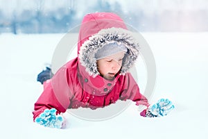 Funny Caucasian smiling girl in warm winter clothes pink jacket playing with snow. Cute child lying on ground during cold winter