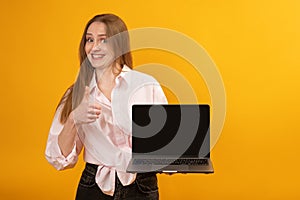 Funny caucasian woman holding laptop computer and shows thumbs up looking at the camera on yellow background. Copy space, mock up