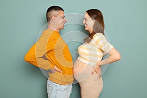 Funny cheerful family waiting baby  over blue background standing with hands on hips measuring bellies expectant parents