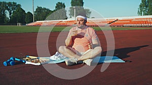 Funny cheerful man after training at the stadium wipes sweat, sits on a mat, eats pizza, drinks beer