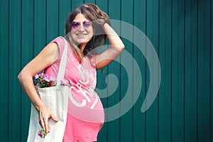 Funny cheerful pregnant woman with eco shopping bag with pink flowers in it on green background. Positive pregnancy, zero waste