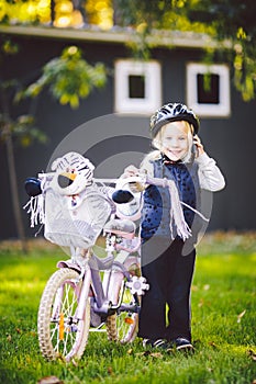 Funny child Caucasian girl blonde in a bicycle helmet near a purple bike with a basket in outside the park on a green