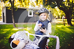 Funny child Caucasian girl blonde in a bicycle helmet near a purple bike with a basket in outside the park on a green lawn grass