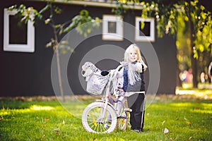 Funny child Caucasian girl blonde near a purple bike with a basket and a zebra toy in an outside park on a green lawn