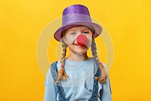 Funny child clown, hat and a red nose. Cheerful little girl on a yellow background. April 1. April Fools Day. Copy space