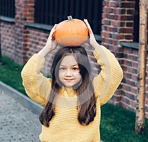 Funny child girl in orange pullover for Halloween with pumpkin and on a dark brick background
