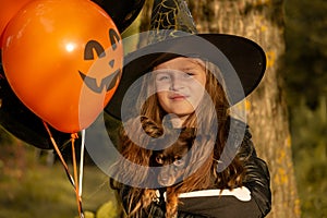 Funny child girl in a witch costume for Halloween with a small dog and with black and orange balloons.