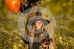 Funny child girl in a witch costume for Halloween with a small dog and with black and orange balloons.