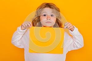 Funny child with sheet of paper, isolated on yellow background. Portrait of a kid holding a blank placard, poster. Empty
