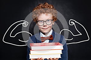 Funny child smart student holding book stack on chalkboard background. Back to school concept