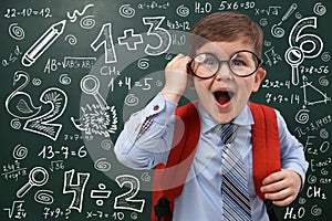 Funny child wearing glasses near chalkboard with mathematical equations. First time at school