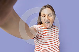 Funny childish woman taking selfie picture, showing tongue out to camera, making front selfportrait.