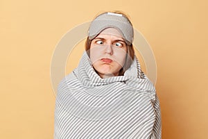 Funny childish woman wrapped blanket and wearing sleeping eye mask isolated over beige background standing with crossed eyes and