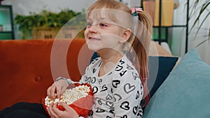 Funny children girl watching comedy video film on tv, eating popcorn on comfortable sofa at home