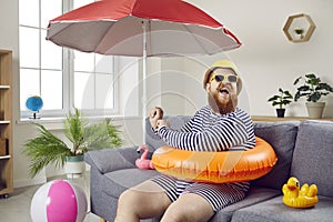 Funny chubby man having fun sitting on sofa in living room with inflatable swimming circle.