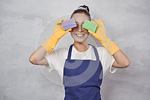 Funny cleaning lady in uniform and rubber gloves covering eyes with kitchen sponges for cleaning and washing dishes