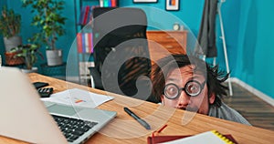 Funny, comical, man with round, thick glasses, disheveled hair, leans out from under desk in
