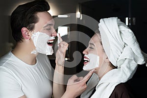 Funny couple beautiful woman applying jokingly foam on handsome man`s nose having fun both with shaving foam on their faces in