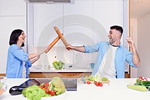 Funny couple fight with baguettes while cooking at home together, husband and wife feeling playful in the kitchen.