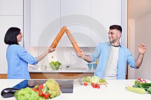 Funny couple fight with baguettes while cooking at home together, husband and wife feeling playful in the kitchen.