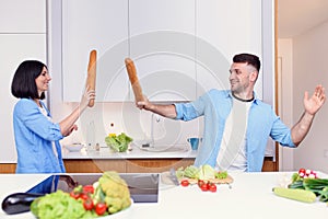 Funny couple fight with baguettes while cooking at home together, husband and wife feeling playful in the kitchen.