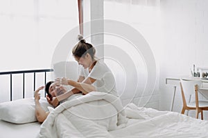 Funny couple wife try to wake lazy husband up in white bedroom.