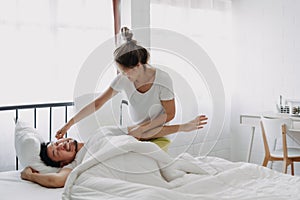 Funny couple wife try to wake lazy husband up in white bedroom.