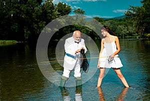 Funny couple of young pretty beauty woman and old fisherman standing in river with fishing rod. Old senior and girl