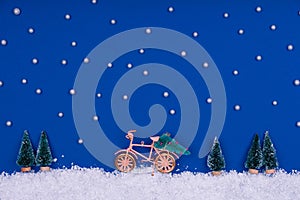 Funny cozy scene with Christmas toys on classic blue. Christmas tree is moved home by bicycle on the snow road with