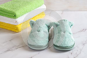 Funny cozy slippers near stack of colored terry towels over marble surface. Concepts of natural cotton toiletries, soft fleece