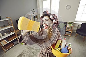 Funny crazy young housewife in curlers and skin care face mask tidying up her house