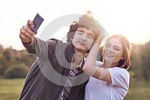 Funny curly male youngster and his girlfriend pose for making selfie portrait against blurred nature background, have positive exp