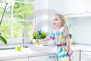 Funny curly toddler girl in colorful dress washing dishes