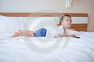 Funny curly toddler lying on the bed with TV remote and attentively watching TV