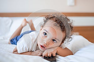 Funny curly toddler lying on the bed with TV remote and attentively watching TV