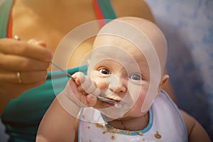 Funny and cute baby eats first meal in a spoon. A newborn boy is smeared with porridge, eats and looks into the camera