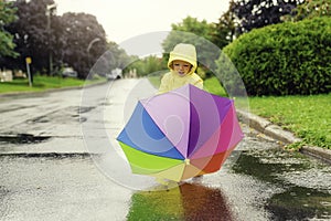 Funny cute baby girl wearing yellow waterproof coat and boots holding colorful umbrella playing in the rain