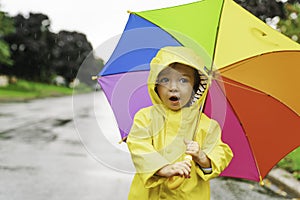 Funny cute baby girl wearing yellow waterproof coat and boots holding colorful umbrella playing in the rain