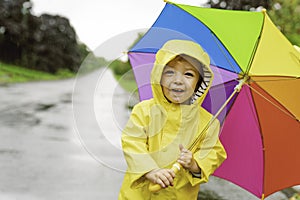 Funny cute baby girl wearing yellow waterproof coat and boots holding colorful umbrella playing in the rain