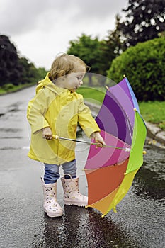 Funny cute baby girl wearing yellow waterproof coat and boots holding colorful umbrella playing in the rain