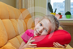 Funny and cute blonde little laughing girl lying on the red pillow on the orange sofa in the children's room.